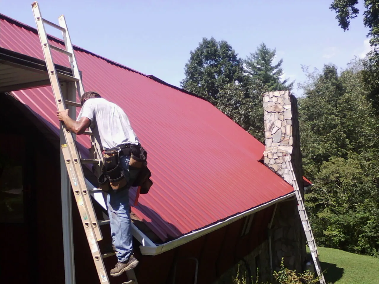 Roofer on a ladder beside a red metal roof and stone chimney — Soffit & Fascia Repair by Roofing Brothers in Queen Anne
